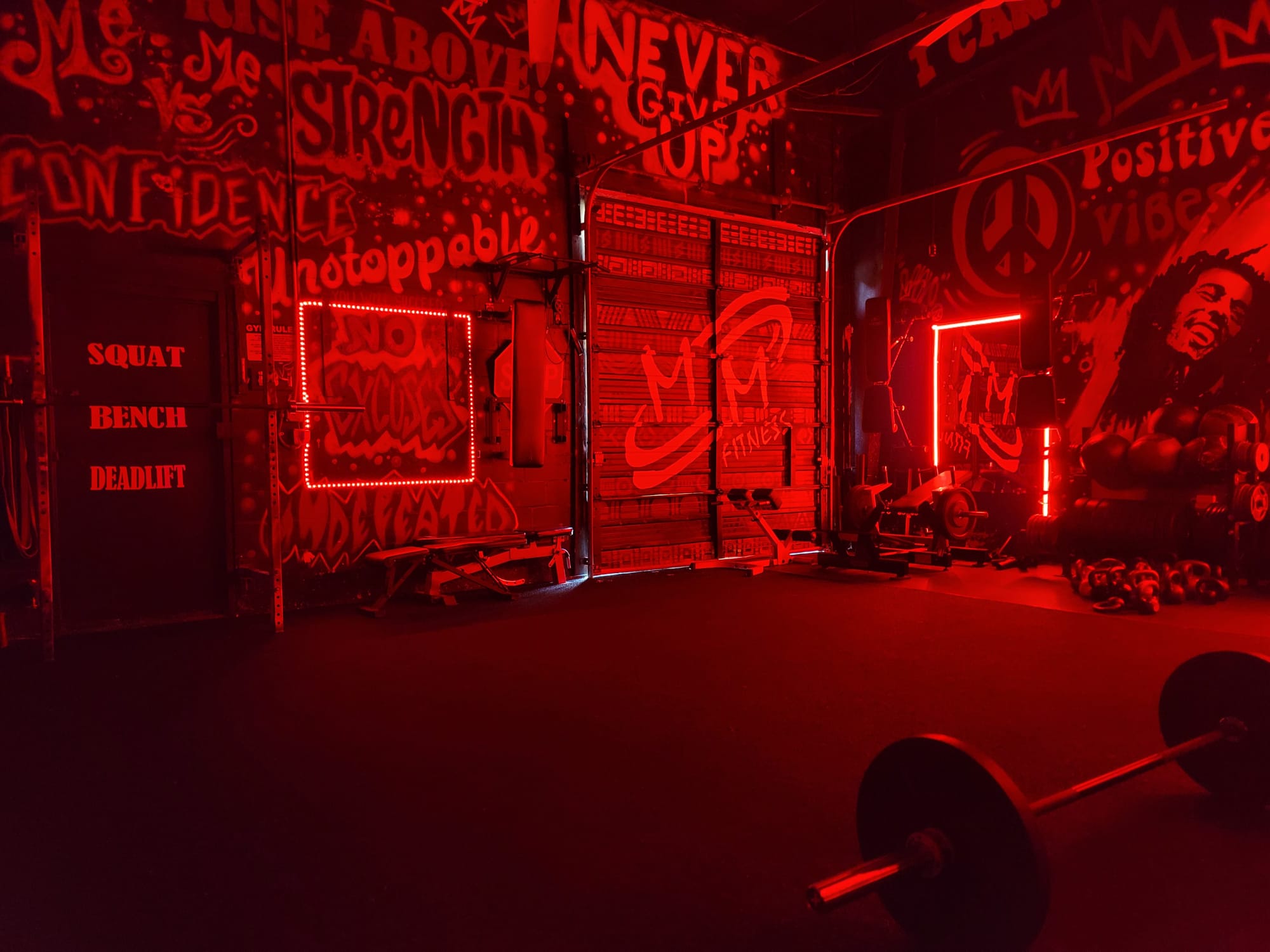 The MMFitness gym floor lit in red, with the MM Fitness logo on the roll-up door and a NO EXCUSES neon sign on the wall
